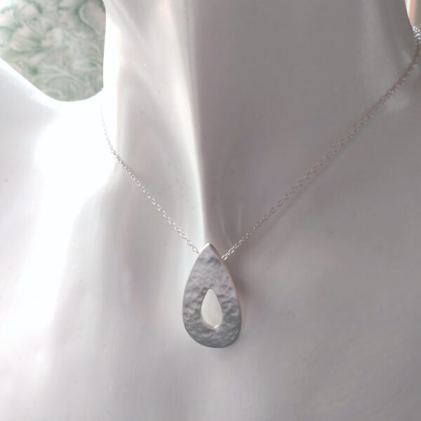 Hammered Silver Teardrop Pendant, in a teardrop shape with a cut out teardrop at the centre. This rustic pendant is handmade in the north-east of England using ethical fine silver. The pendant has a hammered, textured surface and is lightly polished to give a satin finish. The chain goes through a hidden bail on the back of the pendant. UK Hallmarked.