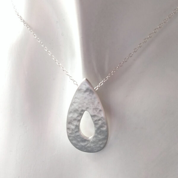 Hammered Silver Teardrop Pendant, in a teardrop shape with a cut out teardrop at the centre. This rustic pendant is handmade in the north-east of England using ethical fine silver. The pendant has a hammered, textured surface and is lightly polished to give a satin finish. The chain goes through a hidden bail on the back of the pendant. UK Hallmarked.