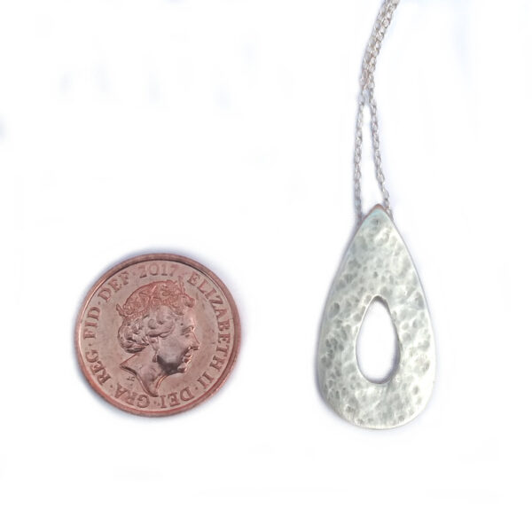 Hammered Silver Teardrop Pendant, in a teardrop shape with a cut out teardrop at the centre. Shown next to a 1 pence piece for size comparison. This rustic pendant is handmade in the north-east of England using ethical fine silver. The pendant has a hammered, textured surface and is lightly polished to give a satin finish. The chain goes through a hidden bail on the back of the pendant. UK Hallmarked.