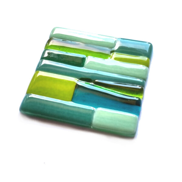 Shades of green fused glass coaster. A small ornamental glass coaster handcrafted using fused glass techniques. The design is abstract irregular green shapes on a clear background. To rest a cup or mug on or to add interest and coloured accents to your room. Handmade in England by Northumbria Gems.