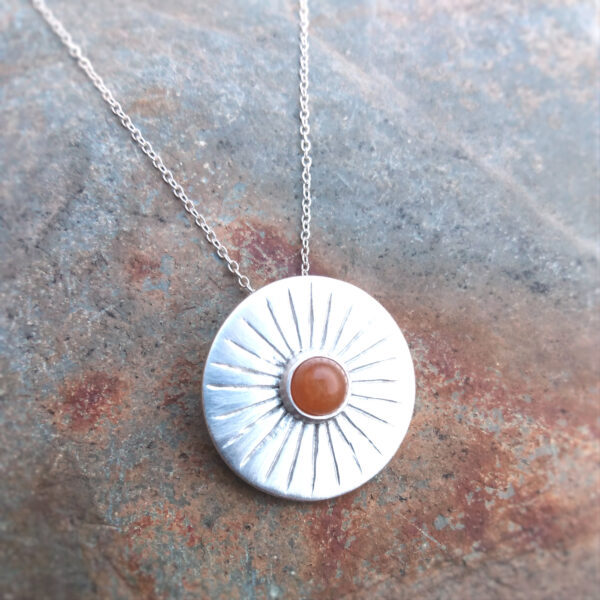 Sun Rays Silver Carnelian Pendant. This round pendant is handcrafted in fine silver. It beautifully captures the essence of sunlight, with delicate silver rays radiating from the central cabochon of natural carnelian. Sourced by hand on the Northumbrian coast, the carnelian gemstone adds a warm, earthy touch to the design. I have given the fine silver a satin finish to complement the pendant design. Handcrafted in England.