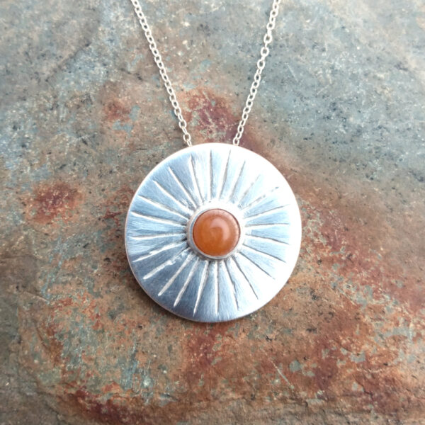 Sun Rays Silver Carnelian Pendant. This round pendant is handcrafted in fine silver. It beautifully captures the essence of sunlight, with delicate silver rays radiating from the central cabochon of natural carnelian. Sourced by hand on the Northumbrian coast, the carnelian gemstone adds a warm, earthy touch to the design. I have given the fine silver a satin finish to complement the pendant design. Handcrafted in England.