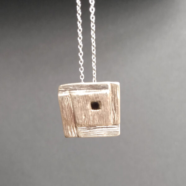 Whitby Jet Inlaid Silver Geometric Pendant. The pendant is square in shape with a stepped, spiral effect design and handcrafted in ethical silver, with a hidden bail at the back. Seen against a plain grey backdrop. The tiny square cabochon has been handcrafted in genuine Whitby jet. Made in the north-east of England.