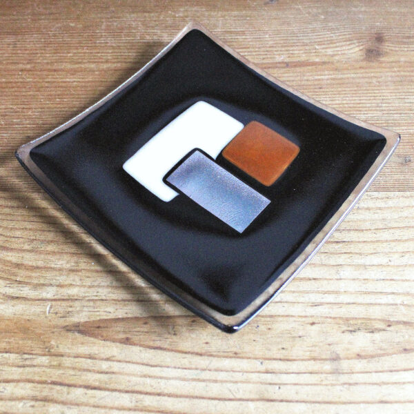 Geometric glass dish in browns and silver on black handcrafted using fused glass techniques. Retro glass dish with tan and peach sections intersected with silver, on a black background.