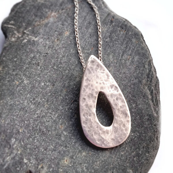 Hammered Silver Teardrop Pendant, in a teardrop shape with a cut out teardrop at the centre. This rustic pendant is handmade in the north-east of England using ethical fine silver. The pendant has a hammered, textured surface and is lightly polished to give a satin finish. The chain goes through a hidden bail on the back of the pendant. UK Hallmarked.